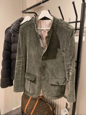 Olivgrön Suede kavaj från Limitato - Snygg olivgrön kavaj i suede från Limitato med fyra knappar framtill och två stora fickor med lock. Insidan har ett unikt mönstrat foder i ljusa toner. Rak passform och axelklaffar ger en cool touch. Perfekt för dig som vill sticka ut lite extra. Storleken är m men den sitter som s-m. Kavajen är i slim fit. Knappen längst ner saknas och det är den ända defekten på kavajen. Modellen är 181 cm. Nypris: 4500.