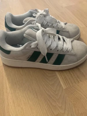 Grå/vita adidas campus med gröna detaljee - Säljer adidas campus i vit/grå mocka. Vita snören och mörkgröna detaljer. Storlek 39. Köpta här på Plick men kom en till användning. 