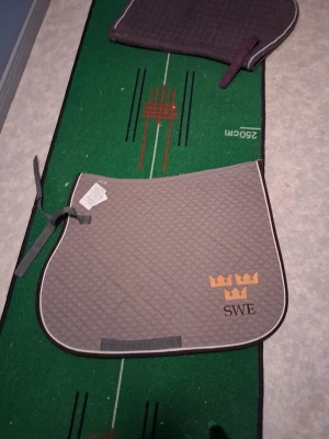 Grått schabrak med SWE och kronor - Säljer ett grått schabrak med rutmönster och guldfärgade kronor samt texten SWE broderat i hörnet. Schabraket har kantband i vitt och grått samt fästen för sadel. Perfekt för dig som gillar stilrena hästtillbehör.  storlek ponny