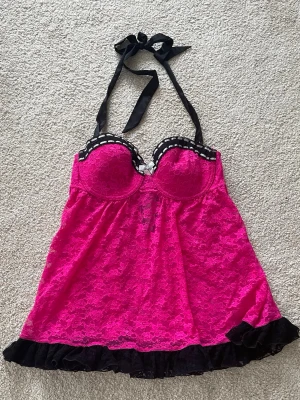 Rosa spetslinnetopp med volang - Snygg rosa linnetopp med svart spetsdetalj och volangkant. Toppen har vadderade kupor, knytband i nacken och dekorativ spets över hela plagget. Perfekt för dig som vill ha en feminin och lekfull look. Passar som S (B kupa ungefär)