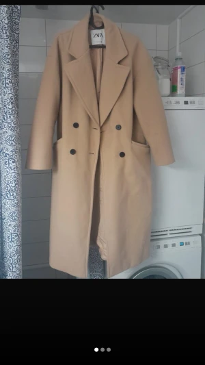 Beige dubbelknäppt rock från ZARA - Stilren beige rock från ZARA med dubbelknäppning och stora slag. Rocken har två framfickor och en rak, lång passform som ger en klassisk look. Perfekt för dig som vill ha en tidlös och trendig ytterrock.