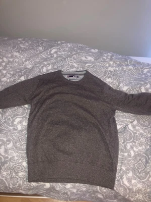 Grå stickad tröja från Morris - Mysig grå stickad tröja från Morris med rund halsringning och diskret broderad logga på bröstet. Tröjan har ribbstickade muddar vid ärmslut och nederkant. Perfekt för lager-på-lager och enkel att matcha med jeans eller chinos.