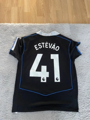 Chelsea FC matchtröja Nike svart - Säljer en svart Chelsea FC matchtröja från Nike med blå detaljer och vit krage. På ryggen står det ESTÊVÃO och nummer 41 i vitt. Tröjan har klubbmärke och Nike-logga på bröstet. Perfekt för fotbollsfans som vill sticka ut på planen.