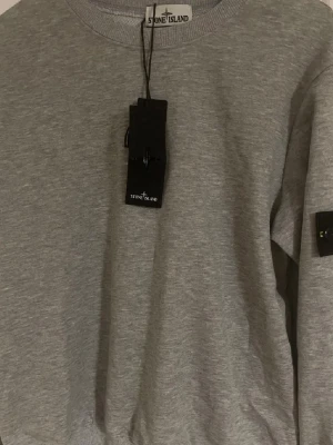 Grå sweatshirt från Stone Island - Snygg grå sweatshirt från Stone Island med klassisk rund hals och ribbade muddar. Tröjan har den ikoniska svarta patchen med gul kompasslogga på ärmen. Perfekt för en stilren och avslappnad look. Kan diskutera pris. 