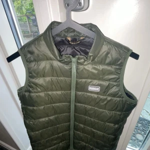 Olivgrön dunväst från Barbour XL - Snygg olivgrön dunväst från Barbour International i storlek XL. Västen har quiltad design, hög krage och dragkedja framtill. Perfekt att slänga över en hoodie för extra värme. Klassisk Barbour-logga på bröstet och lätt vadderad känsla.