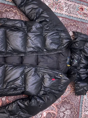 Svart dunjacka från Polo Ralph Lauren - Snygg svart dunjacka från Polo Ralph Lauren med huva och dragkedja. Jackan har flera fickor med dragkedja och den klassiska röda loggan broderad på bröstet. Perfekt för kalla vinterdagar tack vare det tjocka dunfodret.