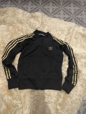 Svart Adidas zip hoodie med guld - Snygg svart zip hoodie från Adidas med klassiska guldiga ränder längs ärmarna och Adidas-logga på bröstet. Tröjan har dragkedja framtill, ribbade muddar och är tillverkad i ett mjukt, sportigt material. Perfekt för en streetwear-look.