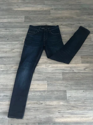 Mörkblå Tiger of Sweden jeans - Säljer ett par mörkblå skinny jeans med klassisk femficksdesign och snygga slitningar vid knäna. Jeansen har en smal passform och är perfekta för en trendig och avslappnad stil. Materialet känns stretchigt och bekvämt.