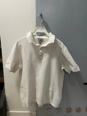 Vit pikéskjorta i bomull - Vit pikéskjorta med korta ärmar och krage. Skjortan har knappar framtill och är tillverkad i ett strukturerat bomullstyg som känns både luftigt och stilrent. Passar perfekt till linnebyxor