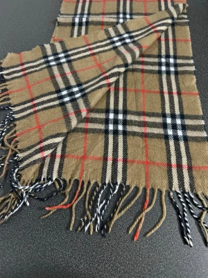 Klassisk rutig halsduk från Burberrys - Säljer en ikonisk halsduk från Burberrys i beige med svart, vitt och rött rutmönster. Halsduken är tillverkad i 100% lammull och har fransar i ändarna. Perfekt accessoar för att lyfta din outfit med en tidlös brittisk stil.