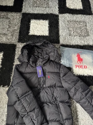 Svart pufferjacka från Polo Ralph Lauren - Svart pufferjacka från Polo Ralph Lauren med huva och dragkedja framtill. Jackan har två sidofickor med dragkedja och en röd broderad logga på bröstet. Perfekt för kalla vinterdagar och riktigt snygg streetstil. Tar också byten.