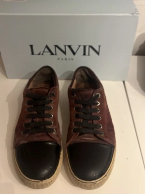 Lanvin bruna sneakers med svart tå - Snygga sneakers från Lanvin i brunt mocka med svart tåparti i läder. Skorna har svarta skosnören och vit sula med retrovibe. Perfekta för dig som gillar exklusiva och stilrena sneakers med en twist. Boxen medföljer.