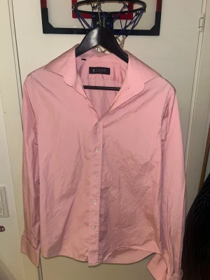 Rosa skjorta från Tiger of Sweden - Säljer en klassisk rosa skjorta från Tiger of Sweden. Skjortan har lång ärm, knappar framtill och en stilren krage. Perfekt för dig som vill ha en enkel men snygg skjorta i garderoben. Materialet är som bomull och passformen är normal.