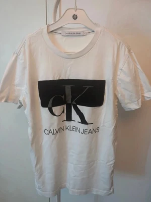  T-shirt från Calvin Klein  - Vit t-shirt från Calvin Klein Jeans med stor svart CK-logga och text på bröstet. Klassisk rund halsringning och korta ärmar. 