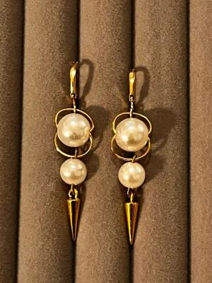 Guldörhängen med pärlor och koner - Säljer ett par unika örhängen i guldfärgad metall med två vita pärlor på varje örhänge. Designen har runda former och avslutas med en spetsig guldfärgad kon. Perfekt för dig som gillar statement-smycken och coola detaljer.