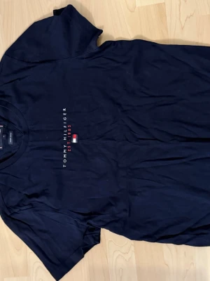 Mörkblå Tommy Hilfiger t-shirt - Snygg mörkblå t-shirt från Tommy Hilfiger med klassisk logga och texten 'EST. 1985' på bröstet. T-shirten har korta ärmar och rund halsringning. Materialet är mjuk bomull och det finns en liten flaggdetalj på ärmen.