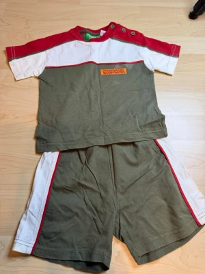 Grönt och rött set från Pulcino Kids - Tvådelat set från Pulcino Kids med t-shirt och shorts i grönt, vitt och rött. T-shirten har knappar vid axeln och en dekorativ etikett framtill. Shortsen har matchande färgblock och röda detaljer längs sidorna. Tillverkat i mjuk bomull, perfekt för varma dagar.