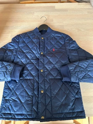 Mörkblå pufferjacka från Ralph Lauren - Snygg mörkblå pufferjacka från Ralph Lauren med quiltat mönster, ribbade muddar och tryckknappar framtill. Jackan har två stora fickor och den klassiska röda loggan broderad på bröstet. Perfekt för kalla vinterdagar och riktigt stilren look.