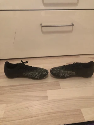 Svarta Puma Future fotbollsskor - Säljer ett par svarta Puma Future fotbollsskor med vita grafiska detaljer på sidorna. Skorna har snörning och låg profil, samt mönstrad ovandel för bättre bollkontroll. Perfekta för dig som vill ha stilrena och funktionella skor på planen.