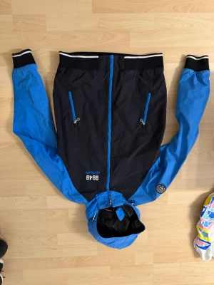 Blå och svart vindjacka 8848 Altitude - Snygg vindjacka från 8848 Altitude i svart med blå ärmar och huva. Jackan har två dragkedjeförsedda fickor fram, ribbade muddar och vit rand på kragen. Materialet är tunt och vindtätt, perfekt för blåsiga dagar. Märkeslogga på bröstet och ärmen.
