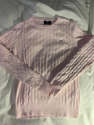 Ljusrosa kabelstickad tröja Gant - Supersöt ljusrosa kabelstickad tröja från Gant i stretchig bomull. Tröjan har rund halsringning, långa ärmar och diskret Gant-logga broderad på bröstet. Perfekt för dig som gillar klassisk och stilren look.