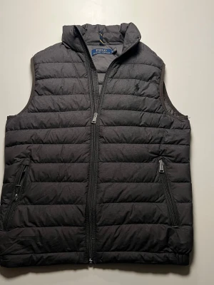  Dunväst från Polo Ralph Lauren - Mörkgrå dunväst från Polo Ralph Lauren med quiltad design och hög krage. Västen har två fickor med dragkedja och diskret logga på bröstet. Perfekt att slänga över en hoodie eller tröja för extra värme.