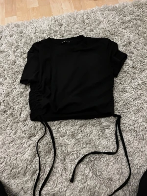 Svart croppad topp med snörning - Snygg svart croppad topp med korta ärmar och snörning i sidorna som ger en cool rynkad effekt. Perfekt att styla med höga jeans eller kjol för en trendig look.