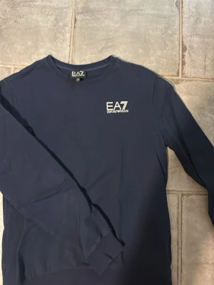 Mörkblå sweatshirt från Emporio Armani - Snygg mörkblå sweatshirt från Emporio Armani EA7 med rund halsringning och diskret logga på bröstet. Tillverkad i mjukt material och har långa ärmar med ribbade muddar. Perfekt för en stilren och avslappnad look.