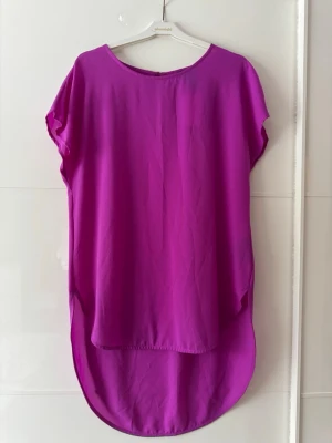 Ny. Lila loose topp med rund hals - Säljer en lila topp med rund halsringning och korta ärmar. Toppen har en loose passform och längre bakstycke för en avslappnad look. Perfekt att styla med jeans eller kjol för en trendig vibe. Förminskad storlek, är som M, bröst 100cm. Lite skavanker, se sista bilden. Köpte så.
