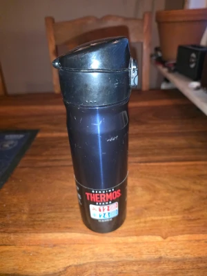 Svart Thermosflaska med lock - Snygg svart thermosflaska från Thermos med praktiskt lock och tryckknapp för enkel öppning. Flaskan är smal och lätt att ta med sig i väskan, perfekt för att hålla drycken varm eller kall under dagen. Tillverkad i rostfritt stål.