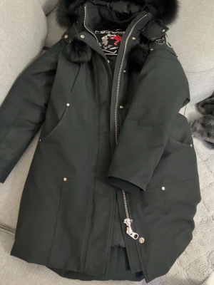 Svart parkas från Moose Knuckles - Snygg svart parkas från Moose Knuckles med stor huva och svart äkta päls. Jackan har silverfärgade detaljer, dragkedja och tryckknappar, samt två stora fickor framtill. Pälsbollar på snörena och logga på axeln ger extra stilpoäng.pris kan diskuteras vid snabb affär 