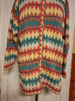 Färgglad retro kofta med zigzagmönster - Unik retro kofta med färgglatt zigzagmönster i turkos, gul, röd, beige och svart. Långärmad modell med knappar framtill och avslappnad passform. Perfekt för dig som gillar vintage vibes och statement-plagg. Stickad i mjukt material.