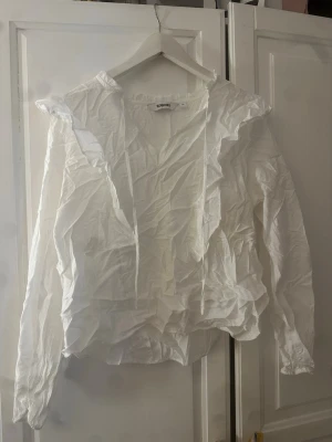 Vit volangblus från Kappahl - Säljer en vit blus från Kappahl med långa ärmar och volangdetaljer vid axlar och halsringning. Blusen har en luftig passform och tunna knytband framtill. Tillverkad i ett lätt och skrynkligt tyg som ger en avslappnad look.