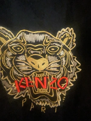 Svart Kenzo t-shirt glittrig  - Säljer en svart t-shirt från Kenzo med ikonisk tigerbrodyr i guld och vitt samt röd Kenzo-text på bröstet. T-shirten har klassisk passform och rund hals. Perfekt för dig som vill sticka ut med en statement-piece från ett välkänt märke.