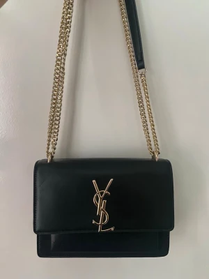 Svart YSL handväska med guldkedja - Snygg svart handväska från Saint Laurent med guldigt YSL-emblem på framsidan. Väskan har en rektangulär form, guldfärgade kedjeremmar och detaljer i svart läder. Perfekt accessoar för dig som gillar lyxig och tidlös design.
