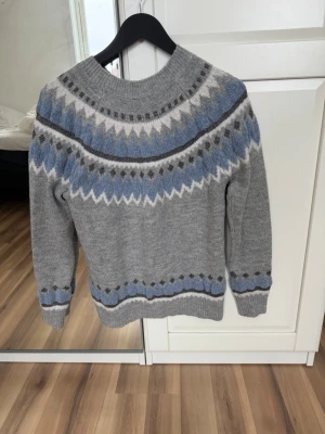 Grå stickad tröja med mönster - Säljer en grå stickad tröja med rund hals och klassiskt nordiskt mönster i blått, vitt och mörkgrått. Tröjan har långa ärmar och ribbade muddar vid ärmslut och nederkant. Perfekt för kalla dagar och riktigt mysig stil.