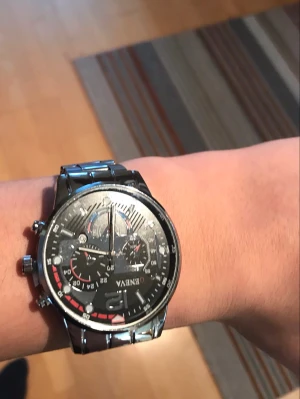 Svart och silverfärgad klocka Genava - Stilren klocka från Genava med svart urtavla och flera små urtavlor för extra funktioner. Klockan har en silverfärgad metall-länk och detaljer i vitt och rött på urtavlan. Perfekt accessoar för dig som gillar en sportig men ändå klassisk look.