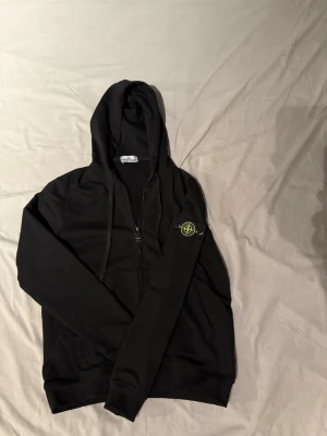 Svart hoodie från Stone Island - Säljer en svart hoodie från Stone Island med dragkedja framtill och klassisk huva med snören. På vänster ärm sitter det ikoniska Stone Island-märket i gult och grönt. Hoodien har ribbade muddar och är tillverkad i mjukt bomullsmaterial.