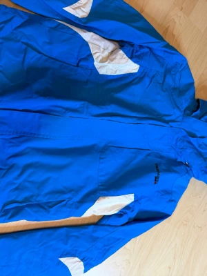 Blå vindjacka från True North - Säljer en blå vindjacka från True North med vita detaljer på ärmar och axlar. Jackan har dragkedja framtill och är tillverkad i ett lätt och tåligt material som skyddar mot vind. Perfekt för dig som vill ha en sportig och stilren look.