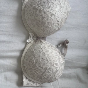 Hunkemöller bh  - Söt vit bh med blommig spets och liten rosett framtill. Bh:n har vadderade kupor och justerbara axelband. Materialet är spets och mesh, vilket ger en romantisk känsla. Perfekt för dig som gillar feminina detaljer.