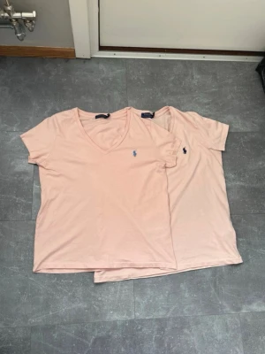 Ljusrosa v-ringad t-shirt Polo Ralph Lauren - Två ljusrosa t-shirts från Polo Ralph Lauren med klassisk v-ringning och blå broderad logga på bröstet. T-shirtsen är kortärmade och gjorda i mjuk bomull, perfekta för en clean och enkel stil.