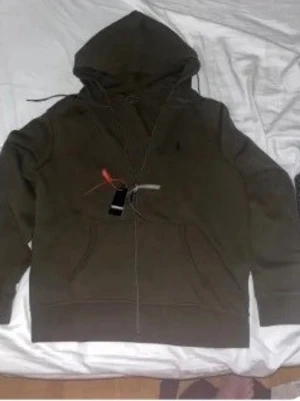 Olivgrön hoodie från Polo Ralph Lauren - Snygg olivgrön hoodie från Polo Ralph Lauren med dragkedja framtill och klassisk broderad logga på bröstet. Hoodien har huva med dragsko och känguruficka. Tillverkad i mjukt bomullsmaterial som känns skönt mot huden. Perfekt för en avslappnad och trendig look.
