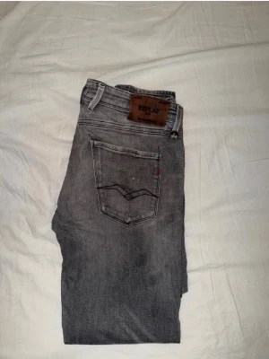 Grå jeans från Replay - Storlek 32/32. Snygga grå jeans från Replay med klassisk femficksdesign och diskreta slitningar. Jeansen har raka ben och detaljerade sömmar på bakfickorna samt en läderpatch med logga i midjan. Tillverkade i mjuk denim för skön passform. Är lite slitna vid skrevet