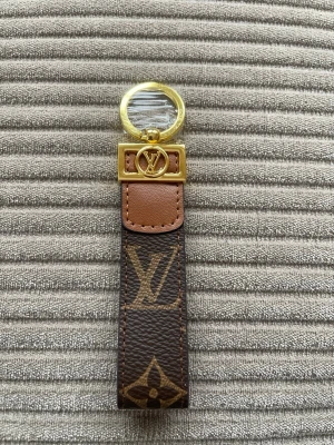  Vuitton nyckelring monogram - Snygg nyckelring från  Vuitton i klassiskt brunt monogrammönster med guldfärgade metalldetaljer och LV-logga. Tillverkad i läder och metall, perfekt accessoar för att ge väskan eller nycklarna en lyxig touch.