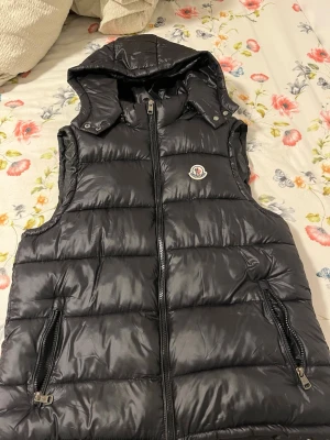 Svart dunväst från Moncler - Snygg svart dunväst från Moncler med avtagbar huva och två dragkedjeförsedda fickor. Västen har quiltad design och Moncler-logga på bröstet. Perfekt för lager-på-lager och håller dig varm under kyliga dagar. Och den har nfc scam den kan scanas.