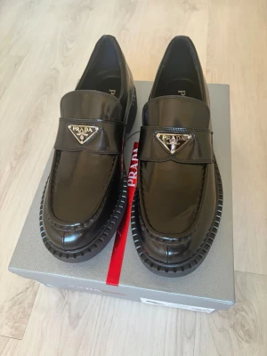 Prada loafers - Snygga svarta loafers från Prada i glansigt läder med chunky sula och ikonisk metallogo framtill. Klassisk design med modern twist, perfekt för dig som vill sticka ut. Skorna har en robust och trendig look med tydliga sömmar och exklusiv känsla.