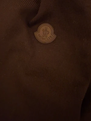 Svart dunjacka från Moncler - Snygg svart dunjacka från Moncler med klassisk logotyp på bröstet. Jackan har hög krage och är tillverkad i ett glansigt material med vaddering som håller dig varm under kalla dagar. Perfekt för vintern och riktigt stilren.