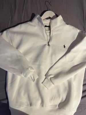 Vit quarter zip från Ralph Lauren - Säljer en stilren vit quarter zip från Ralph Lauren med klassisk broderad logga på bröstet. Tröjan har hög krage med dragkedja och ribbade muddar vid ärmslut och nederkant. Perfekt för en clean och avslappnad look.