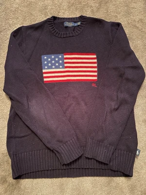 Tröja Polo Ralph Lauren - Den ikoniska mörkblå Polo Ralph Lauren tröjan med den amerikanska flaggan på bröstet. Tröjan har rund halsringning, ribbade muddar och RL-broderiet på magen. Perfekt för dig som vill ha en tidlös tröja!