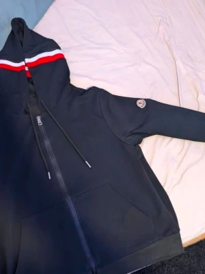 Svart Moncler hoodie med dragkedja - Säljer en svart hoodie från Moncler med dragkedja framtill och fickor. Huvan har röda och vita ränder och dragsnören. Moncler-logga på ärmen ger en exklusiv touch. Perfekt för dig som vill ha en stilren och sportig look.